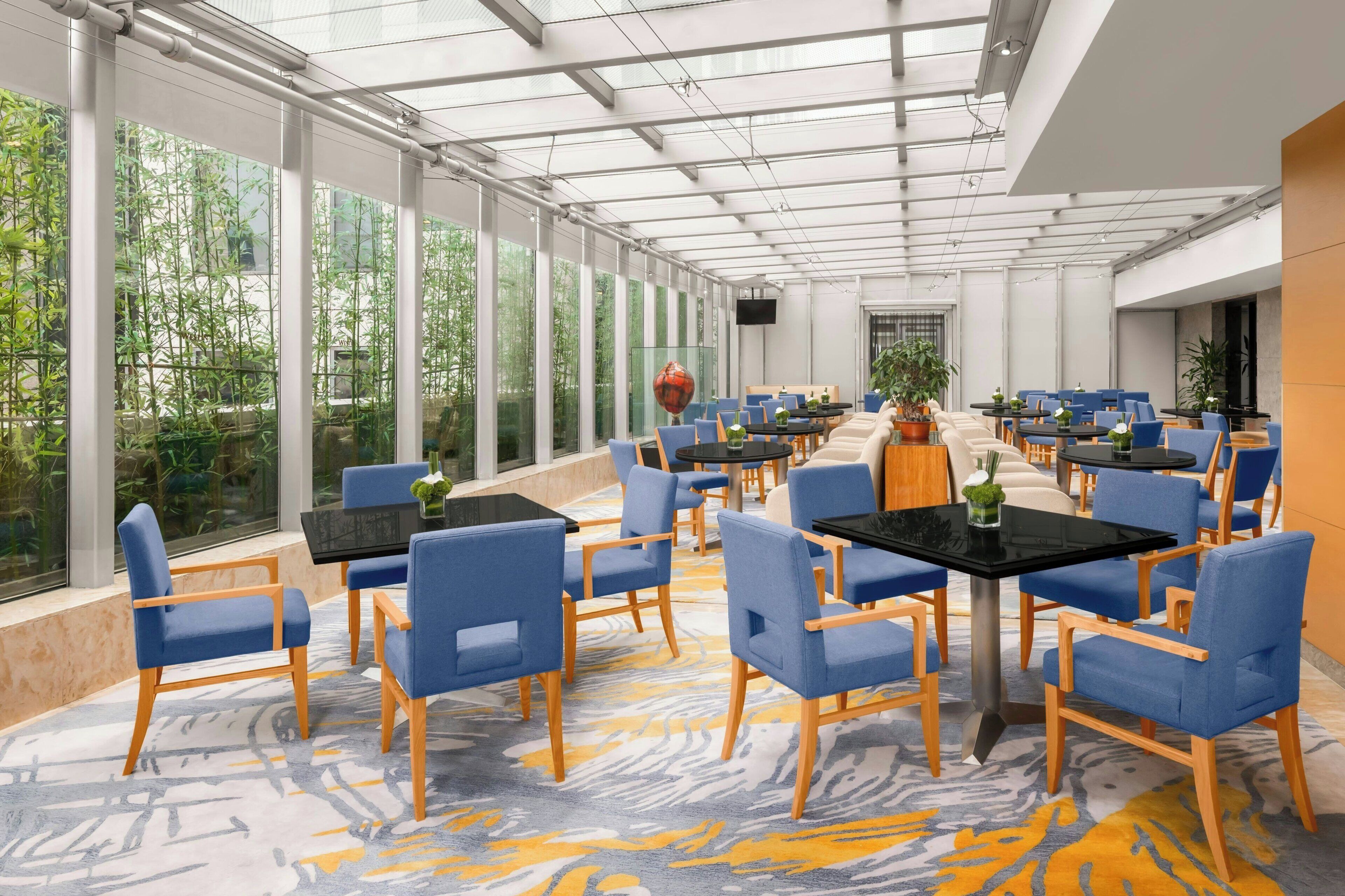 Un espace intérieur vaste et lumineux, avec de grandes fenêtres, des chaises bleues, des tables noires et un tapis à motifs.