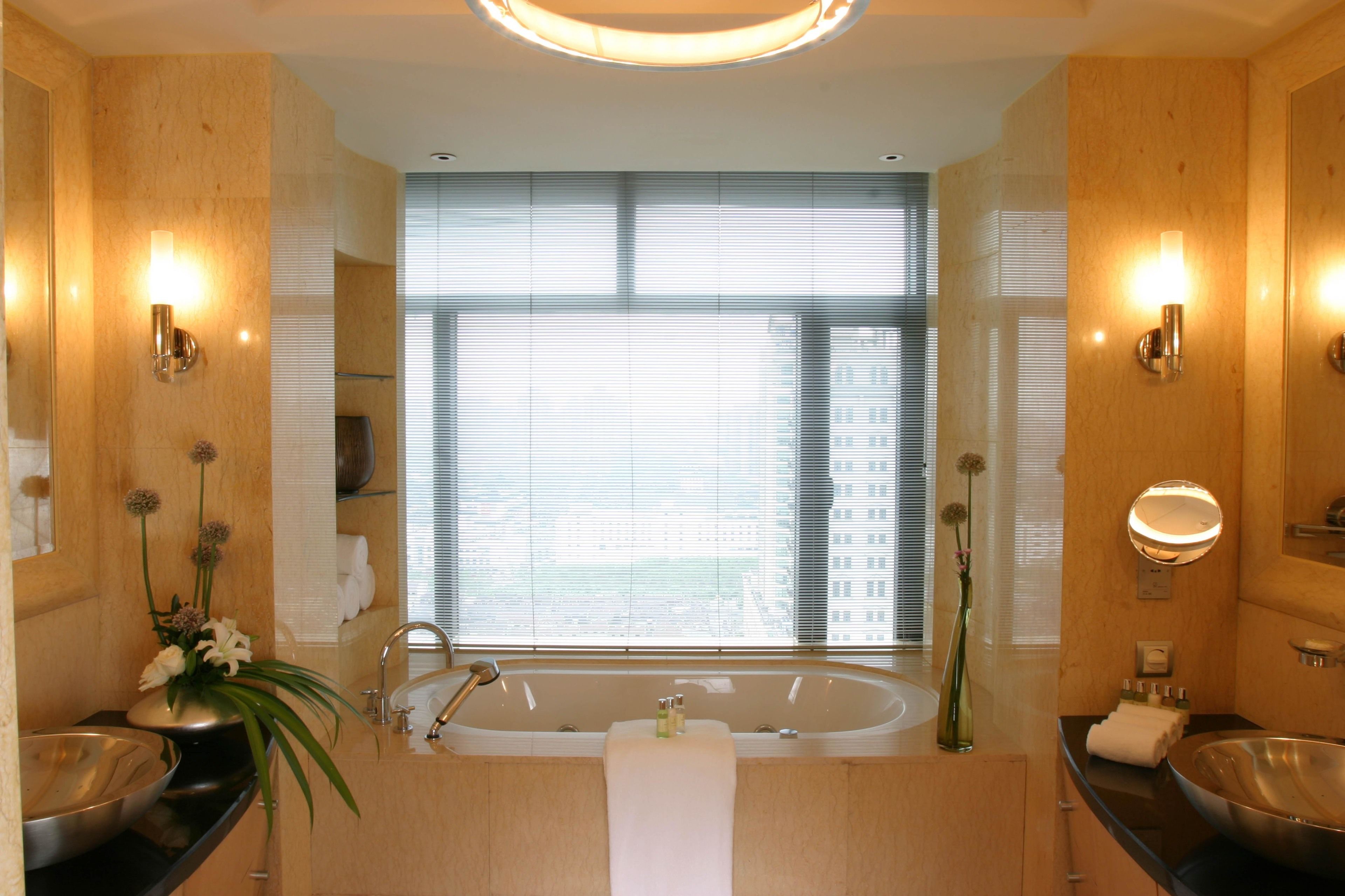 Een moderne badkamer met een groot bad, twee wastafels en een groot raam met jaloezieën.