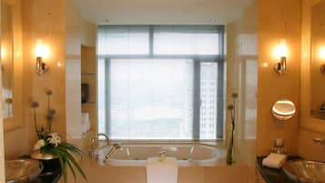 Een moderne badkamer met een groot bad, twee wastafels en een groot raam met jaloezieën.