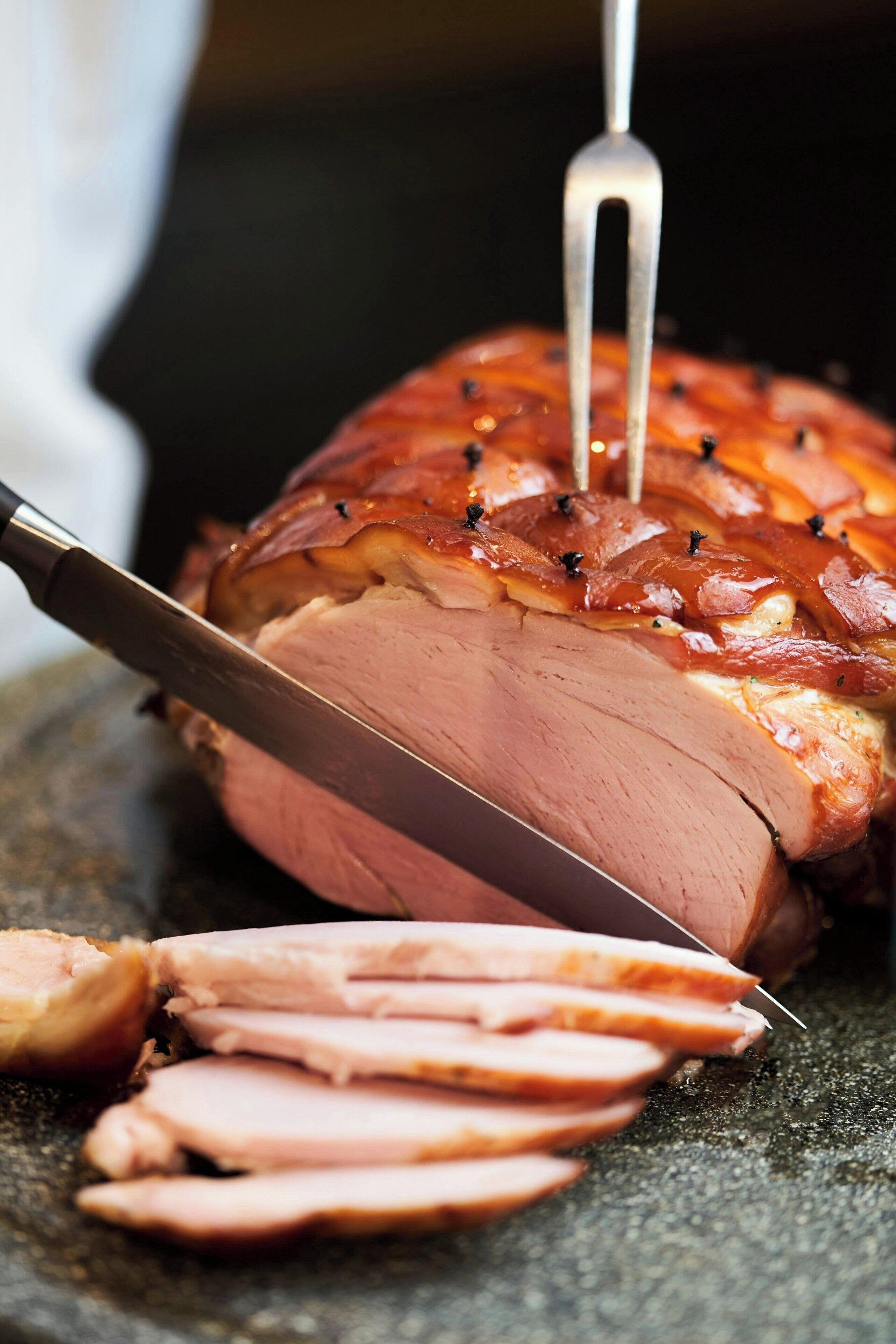 A roasted ham with a knife and fork inserted into it, placed on a dark surface.