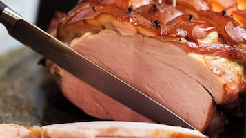 A roasted ham with a knife and fork inserted into it, placed on a dark surface.