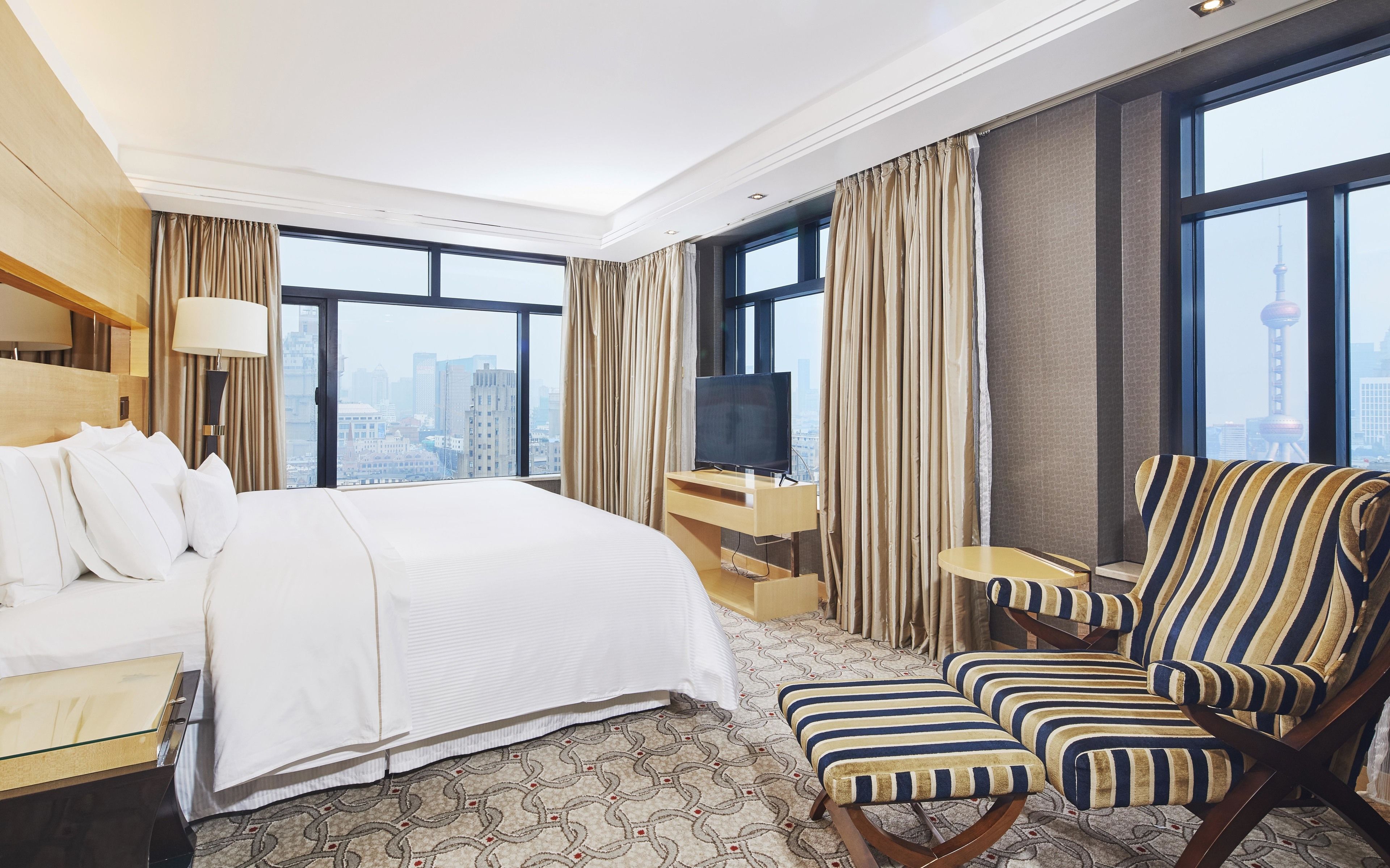 A hotel room with a bed, a TV on a desk, a striped armchair, and large windows with a city view.