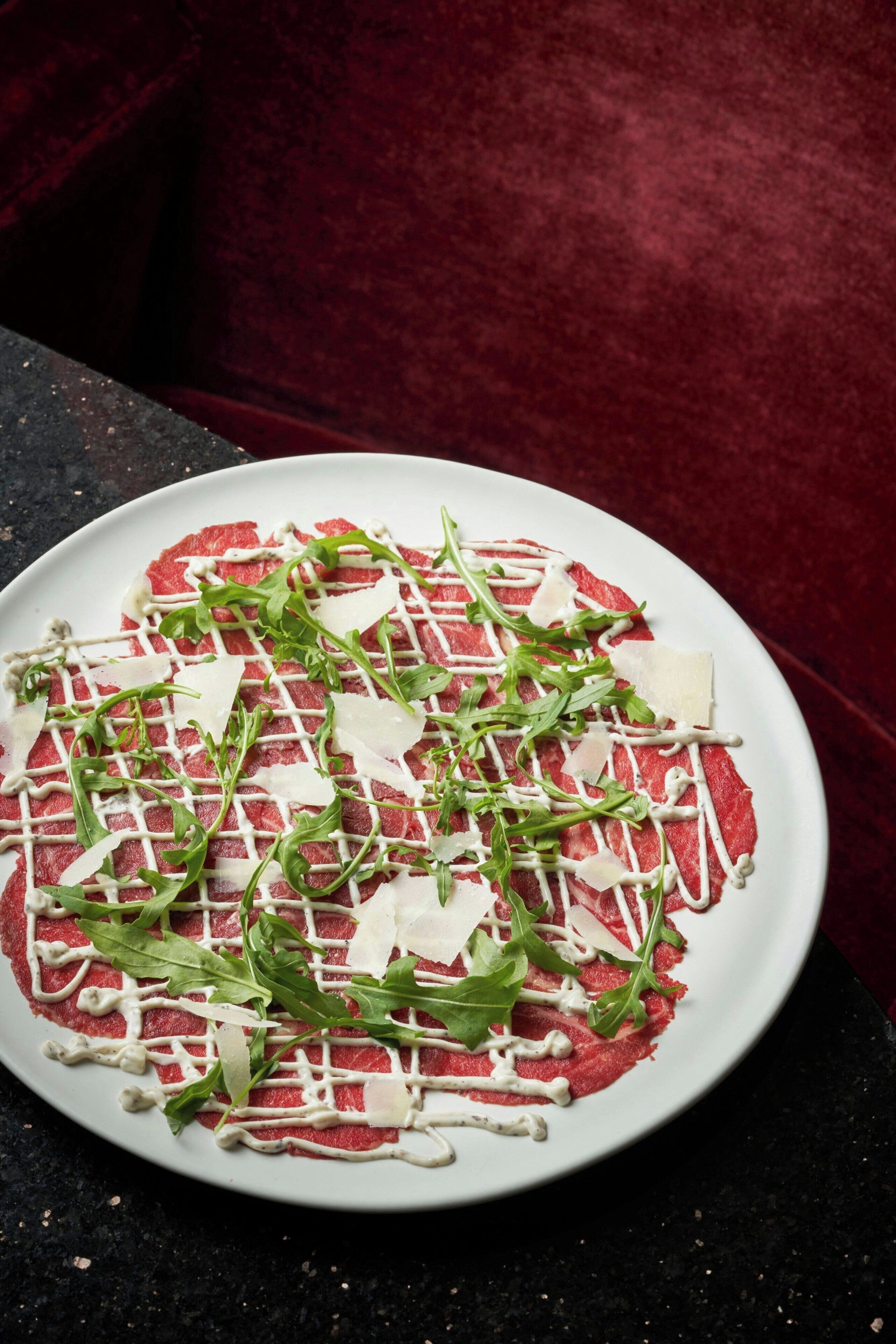 A plate of raw meat topped with greens and cheese, drizzled with sauce, on a dark surface.