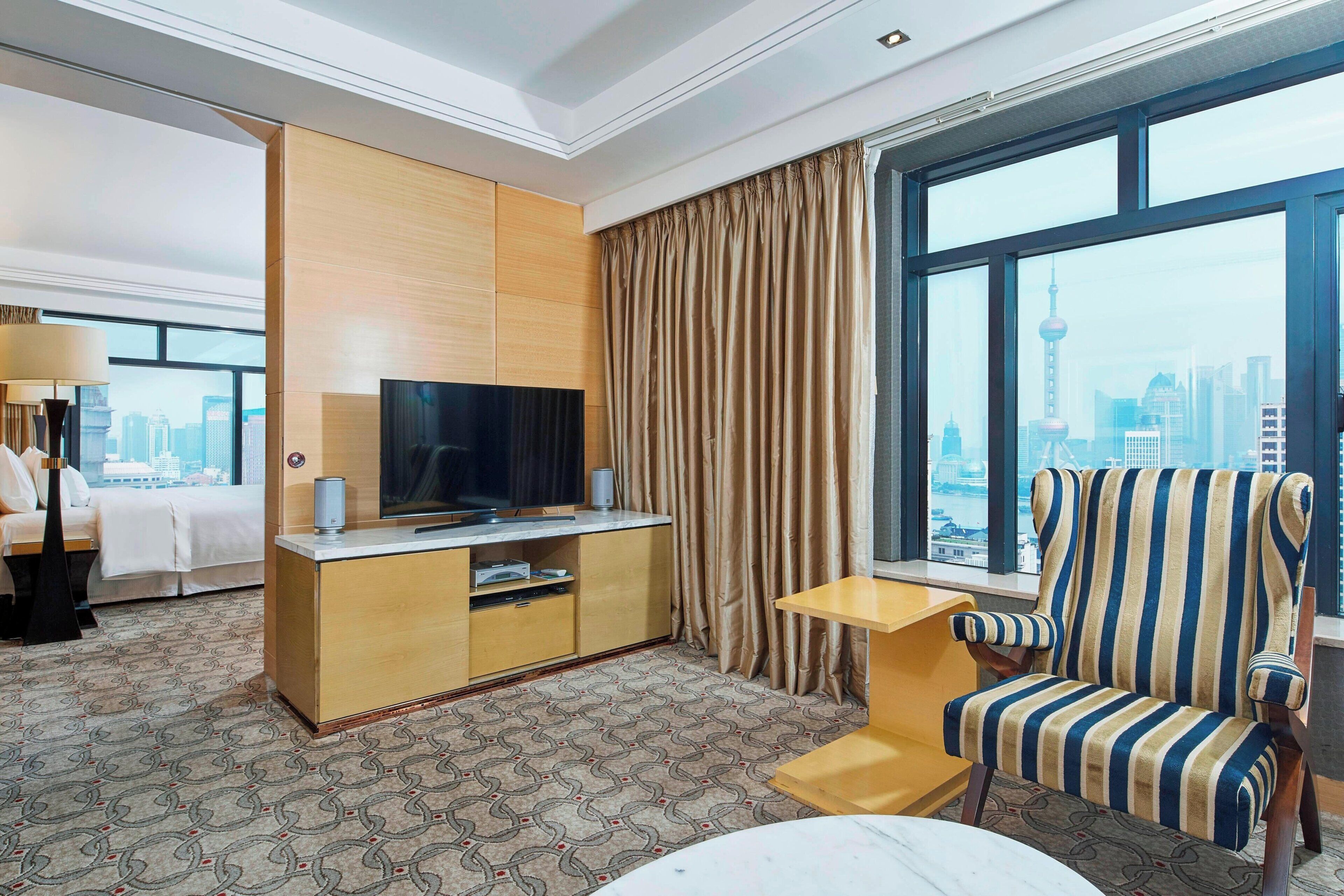 A hotel room with a bed, a TV, a desk, a chair, and a large window with a city view.