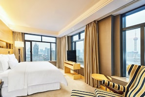 A hotel room with a large bed, a desk with a TV, a chair, and a view of a cityscape.