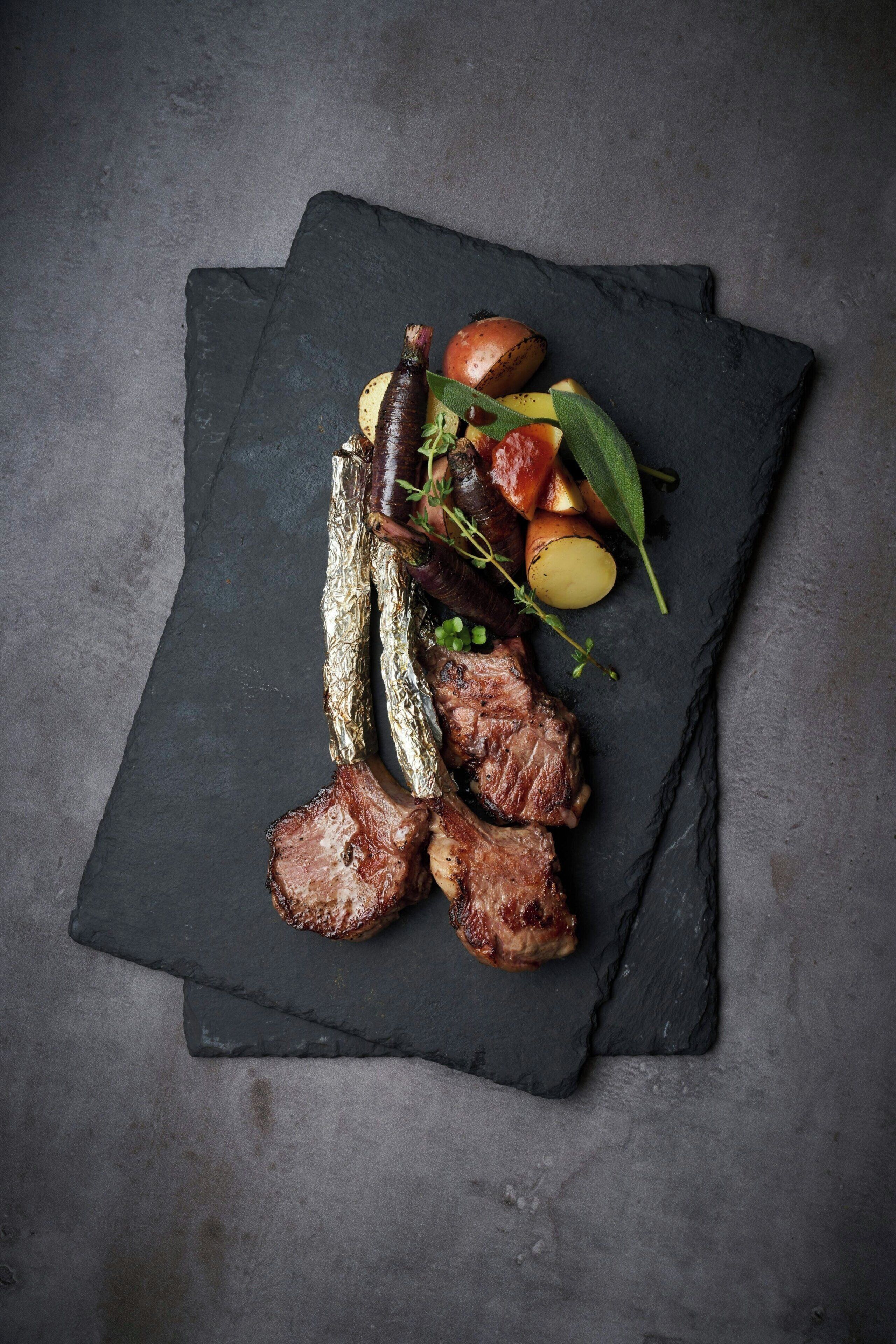 A black slate serving board with grilled meat, potatoes, and herbs.