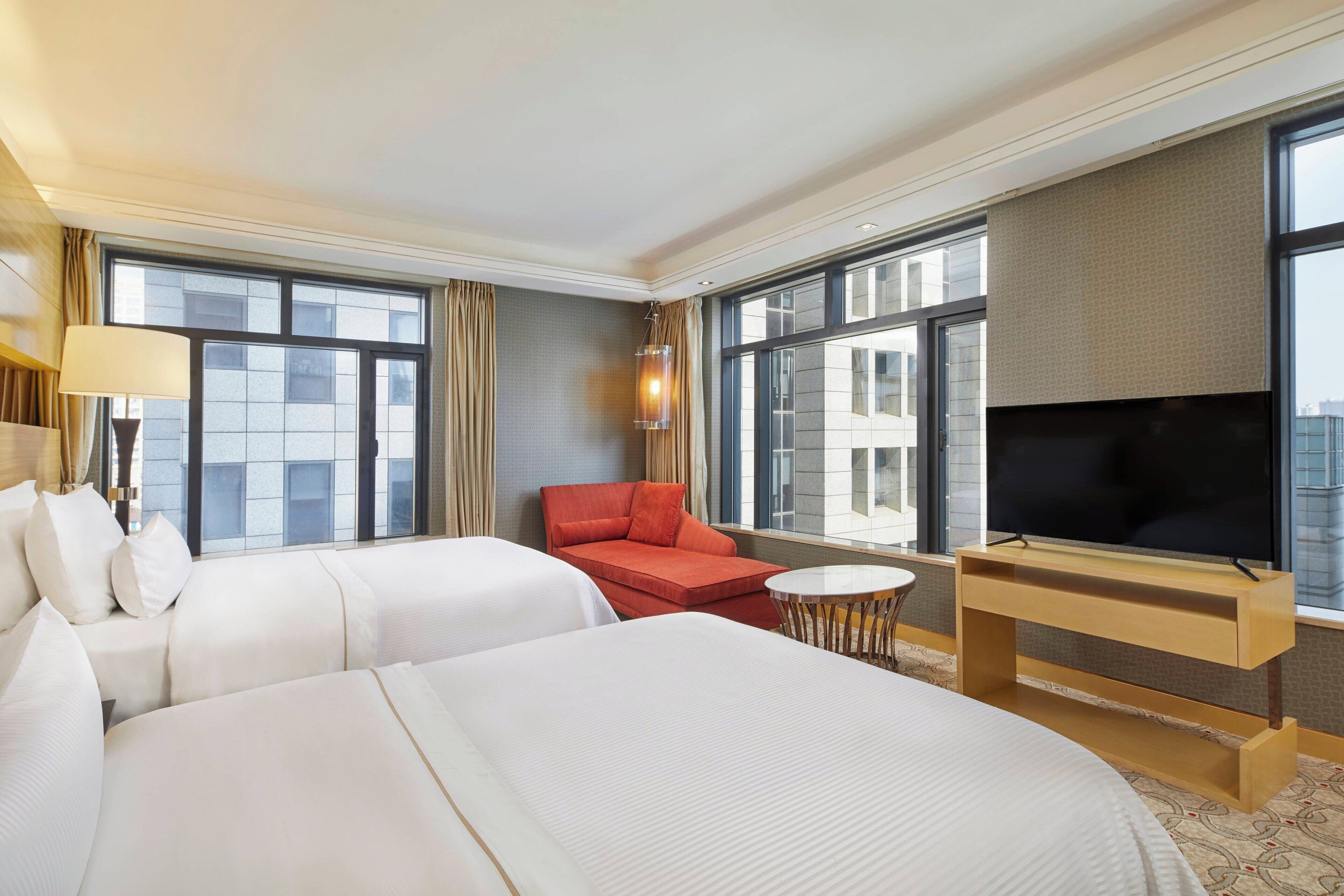 A hotel room with two beds, a red sofa, a TV on a wooden stand, and large windows offering a city view.