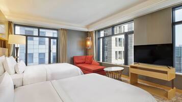 A hotel room with two beds, a red sofa, a TV on a wooden stand, and large windows offering a city view.