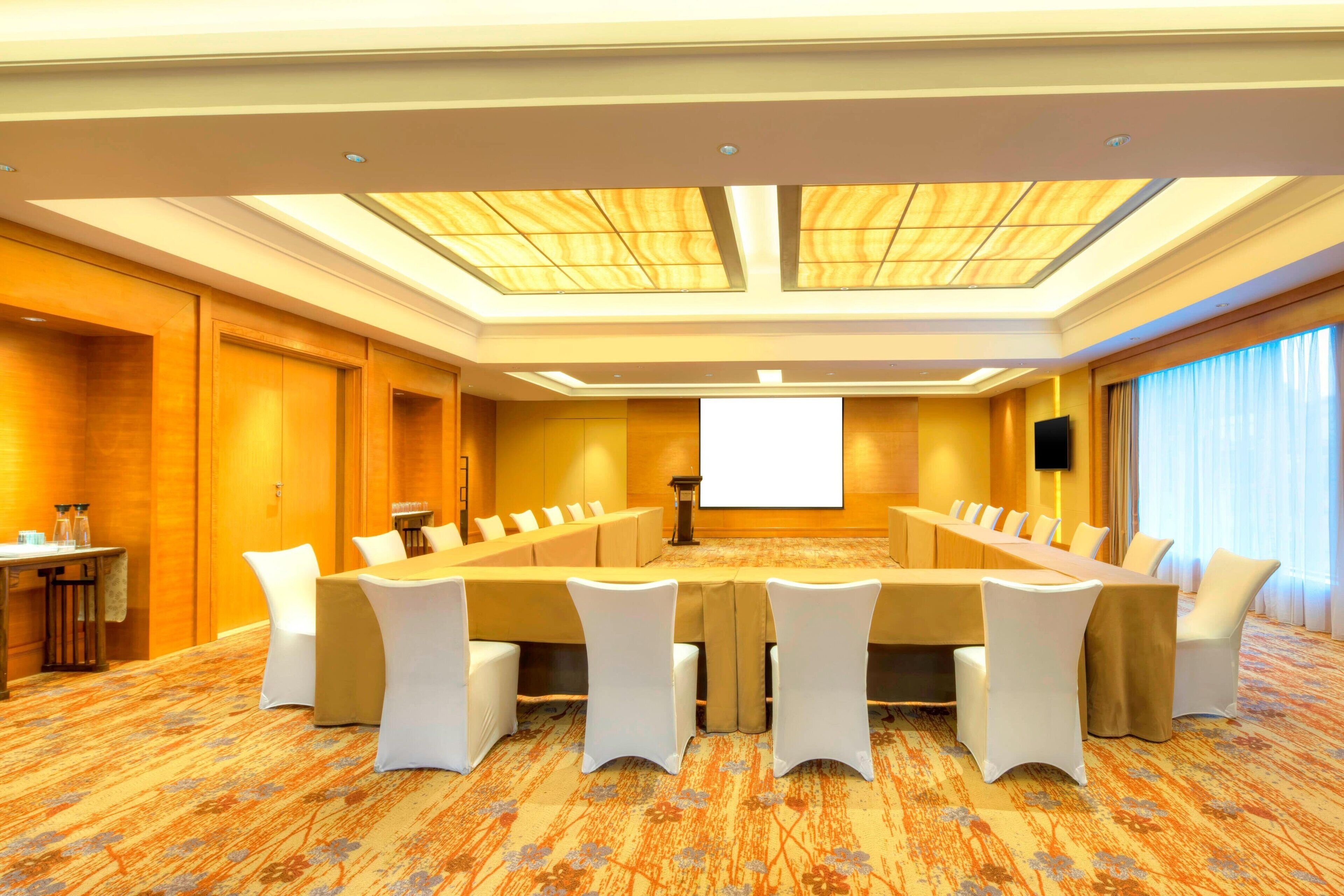 A conference room with a long table, white chairs, a projector screen, and a carpeted floor.