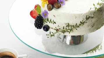 Un gâteau décoré de fruits, présenté sur un présentoir à gâteaux en verre, une tasse de café et une assiette avec une tranche de fruit.