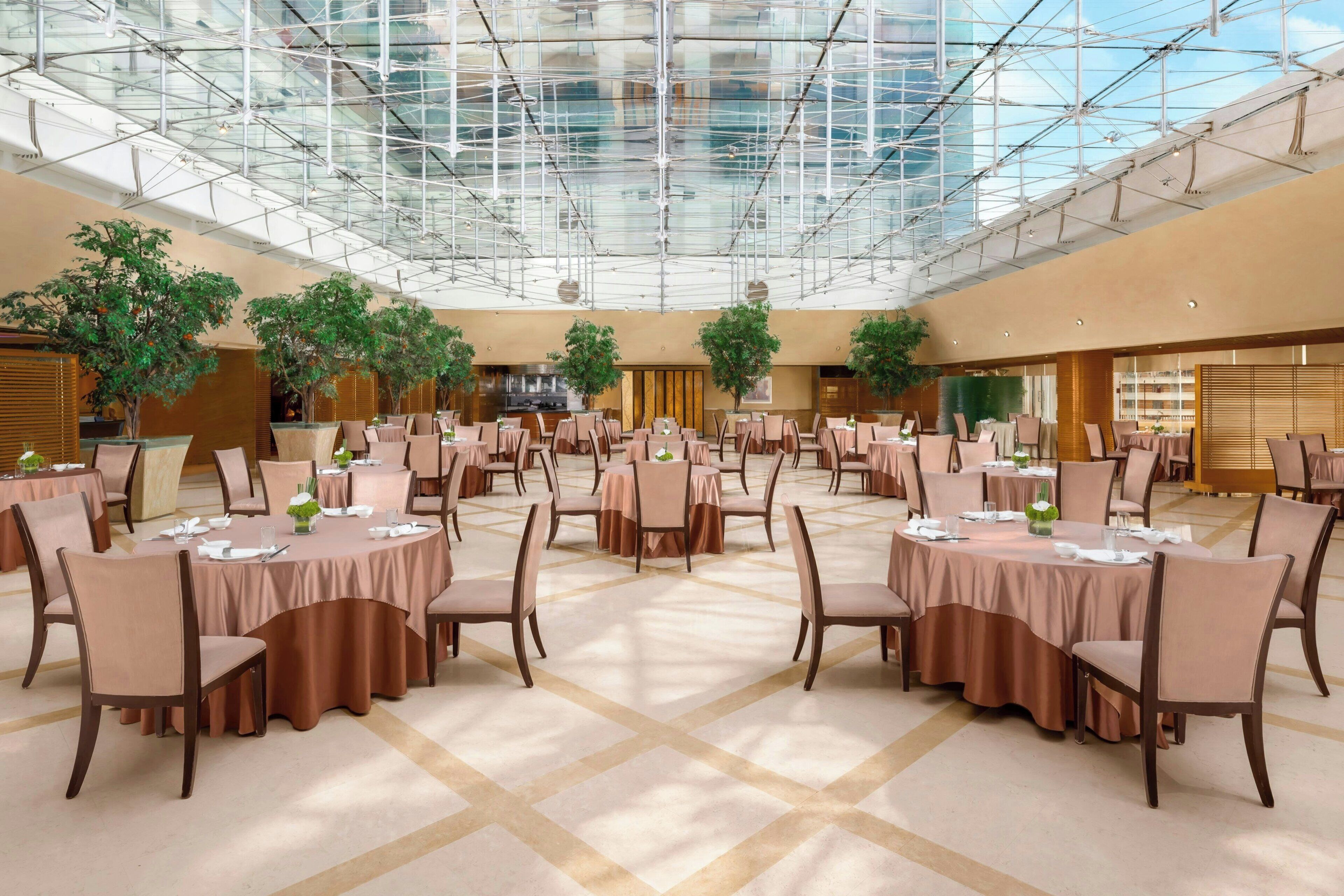 A spacious indoor venue with multiple round tables and chairs, a high ceiling with a glass structure, and potted plants.