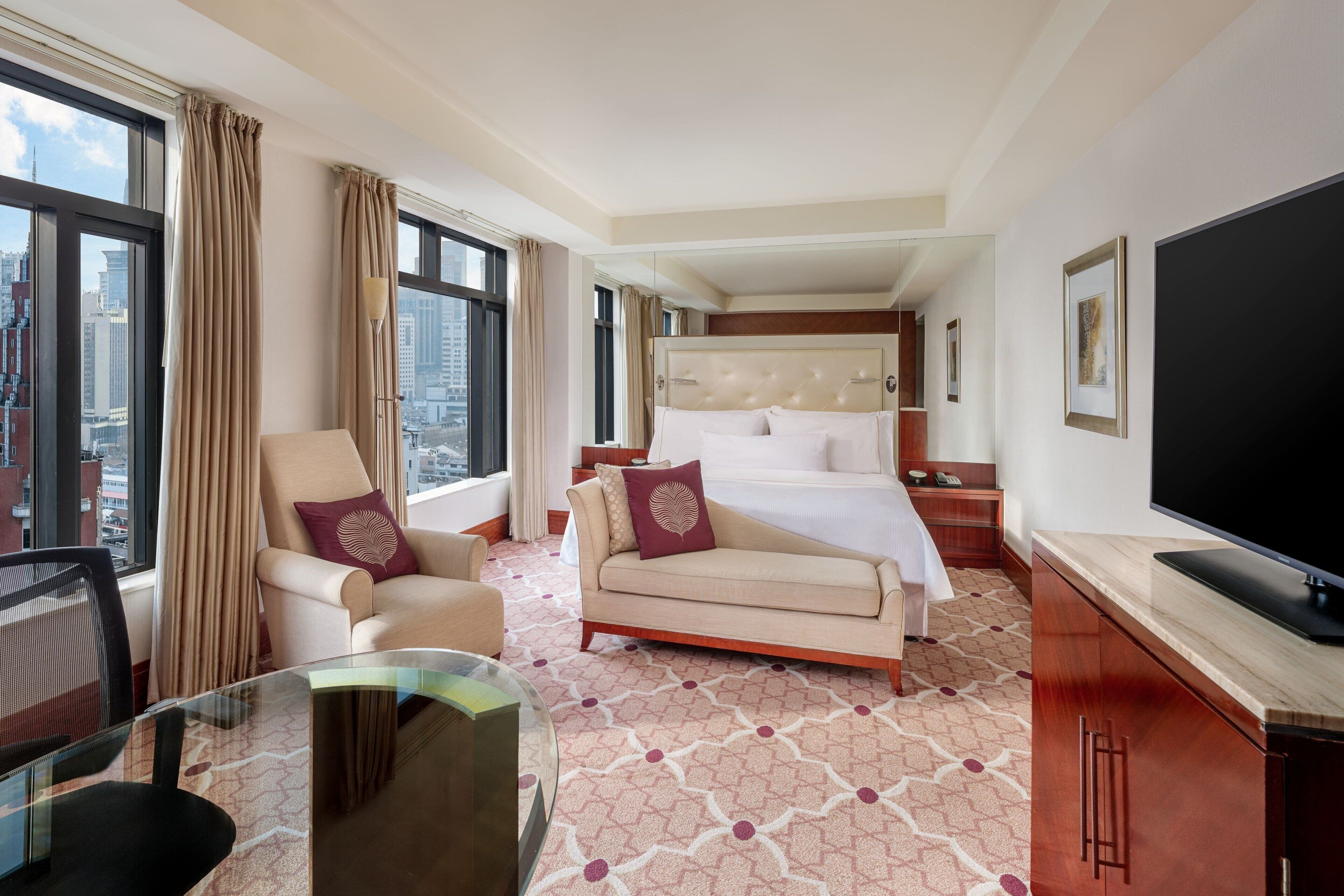 A hotel room with a bed, a sofa, a TV, a desk with a chair, and large windows with curtains.