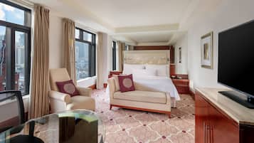 A hotel room with a bed, a sofa, a TV, a desk with a chair, and large windows with curtains.