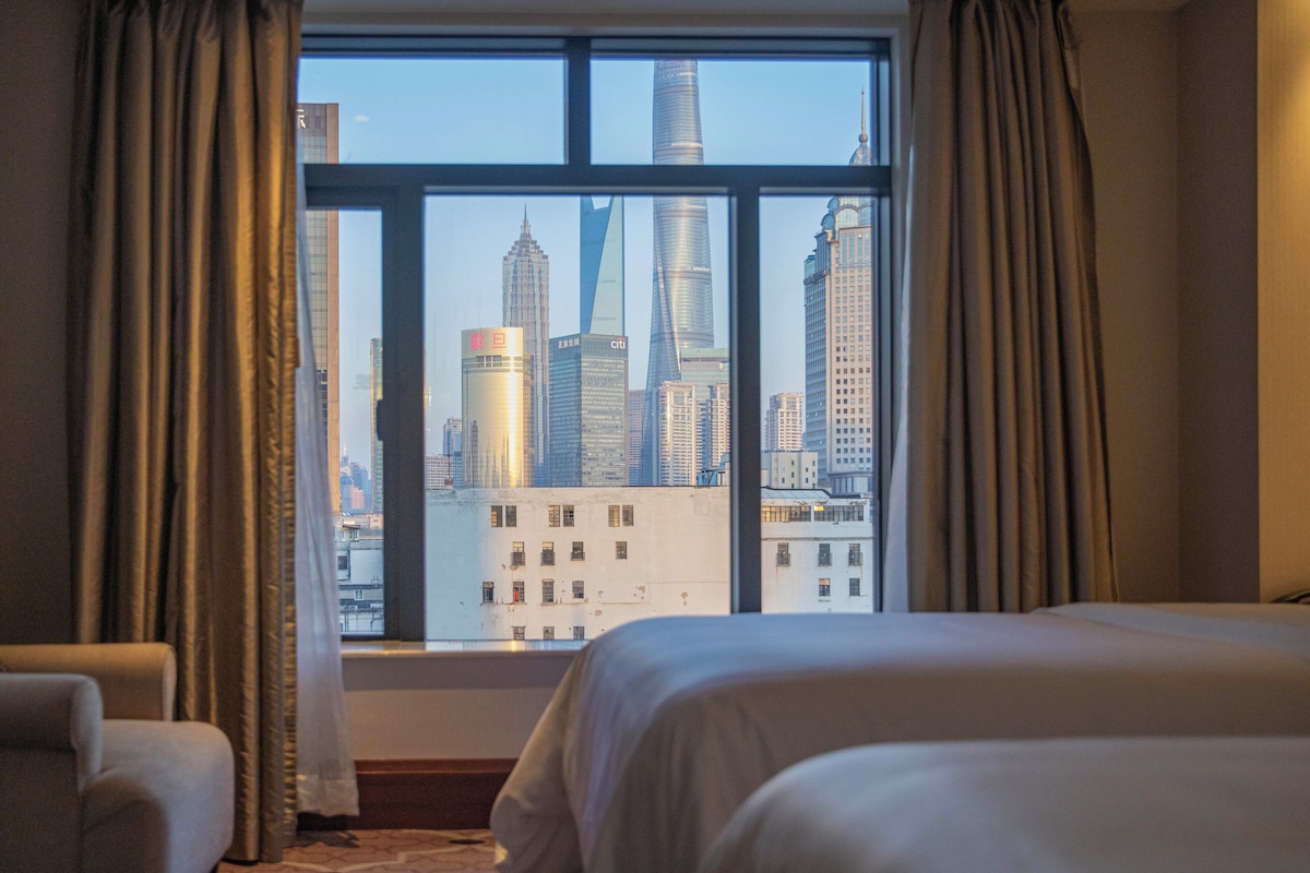 A hotel room with a large window offering a view of a city skyline, including a distinctive skyscraper.