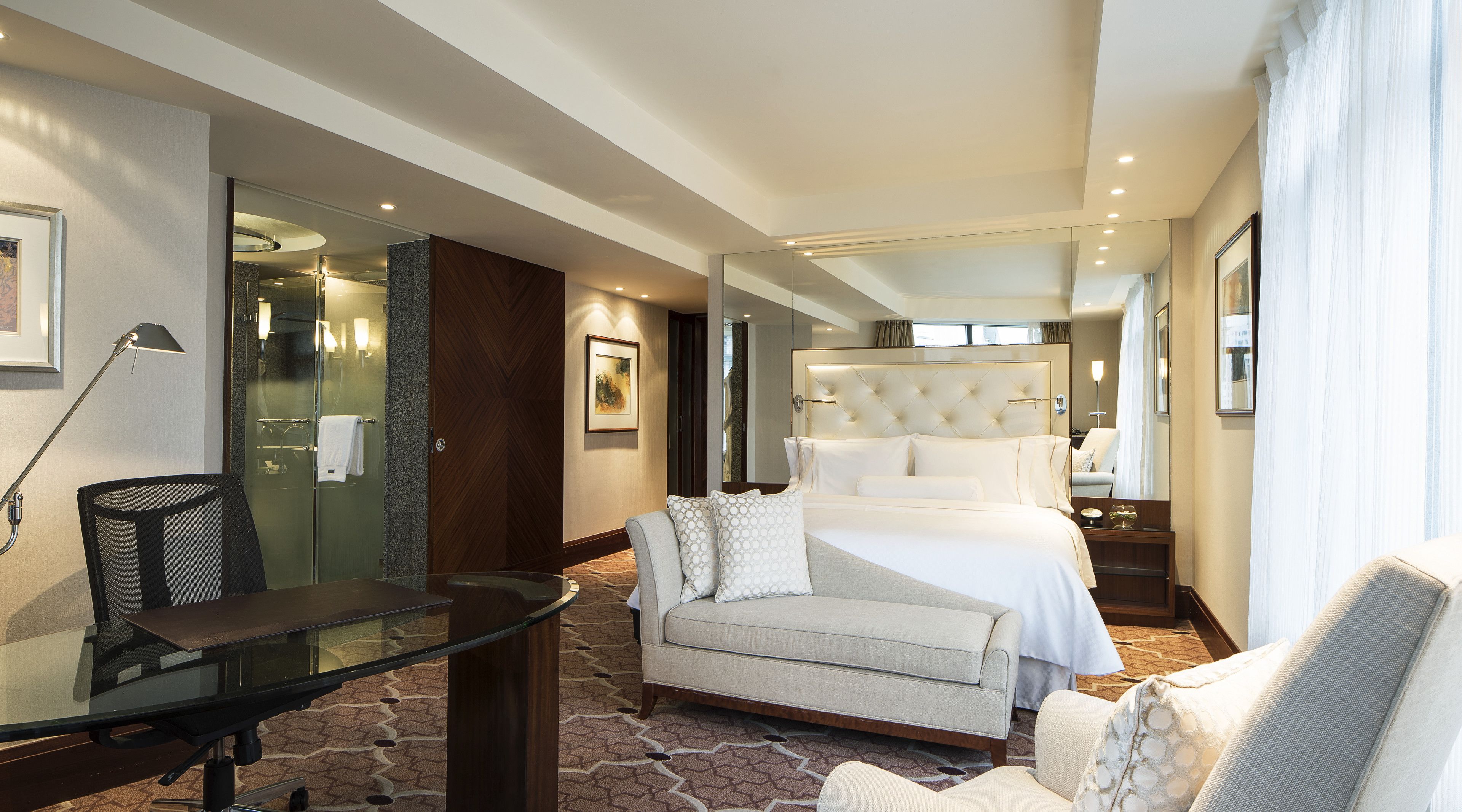 A hotel room with a bed, a sofa, a glass table, and a chair.