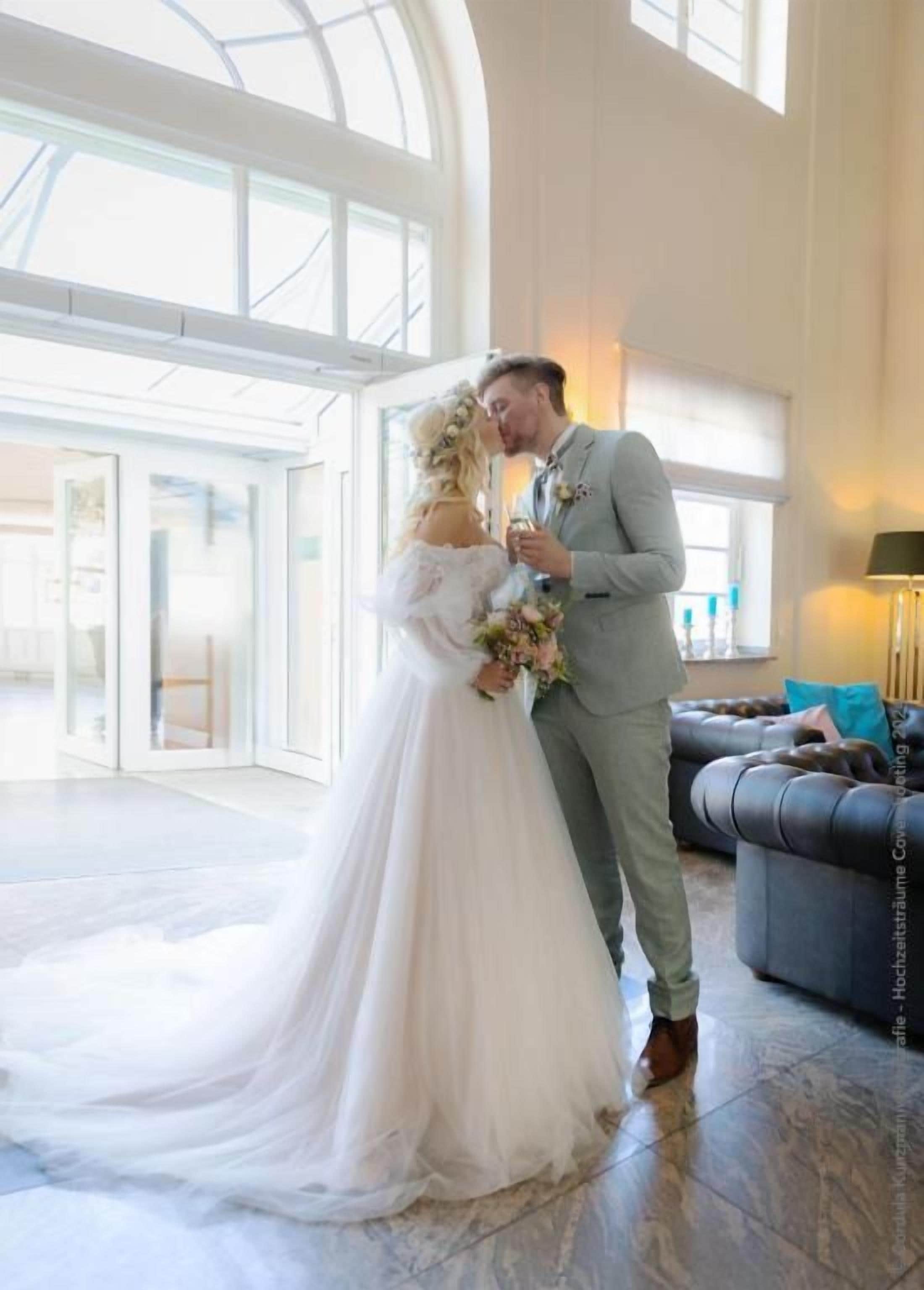 A couple in wedding attire in a spacious room with a large window, a sofa, and a lamp.