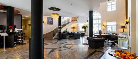 A spacious lobby with a grand staircase, a reception desk, and a seating area with a fruit basket and glasses.