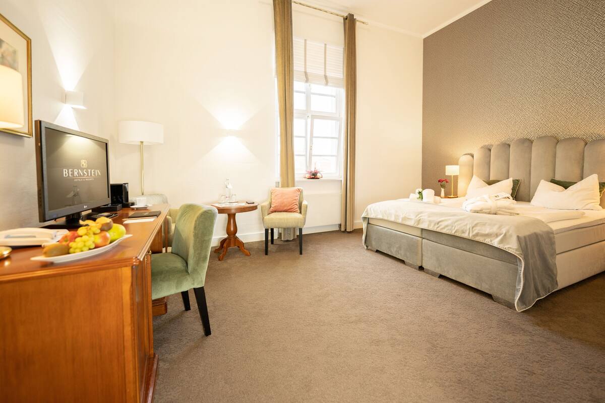 Une chambre d’hôtel comprenant un lit, un bureau avec un ordinateur, une chaise, une petite table avec une lampe et une fenêtre avec des rideaux.