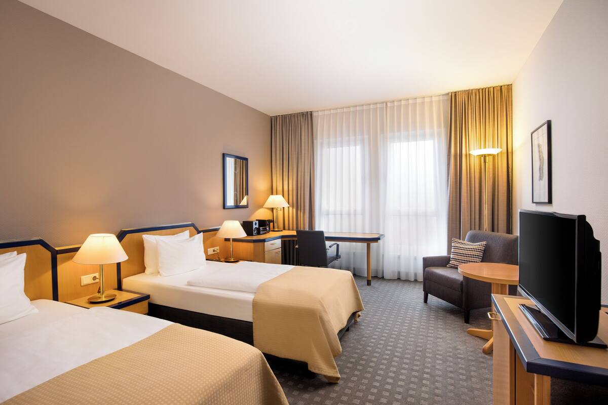Une chambre d’hôtel avec deux lits, un bureau, une chaise, une télévision et une grande fenêtre avec des rideaux.