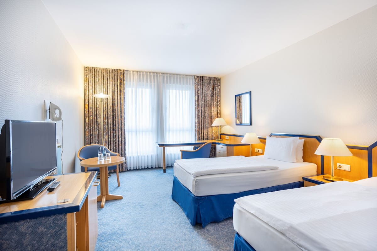 Une chambre d’hôtel avec deux lits, un bureau avec un ordinateur, une télévision et une petite table avec des chaises.