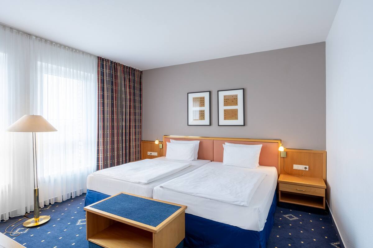 Une chambre d’hôtel avec deux lits, une petite table, une table de chevet et une lampe fixée au mur.