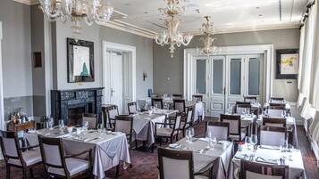 A formal dining room with multiple tables, chairs, chandeliers, and a fireplace.