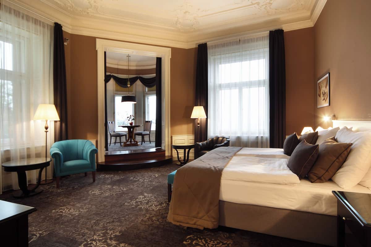 A hotel room with a large bed, two bedside tables, a desk, a chair, and a view of the dining area through an open door.