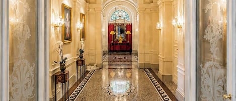 Un amplio pasillo con piso de mármol, decoraciones ornamentales en las paredes y un candelabro.
