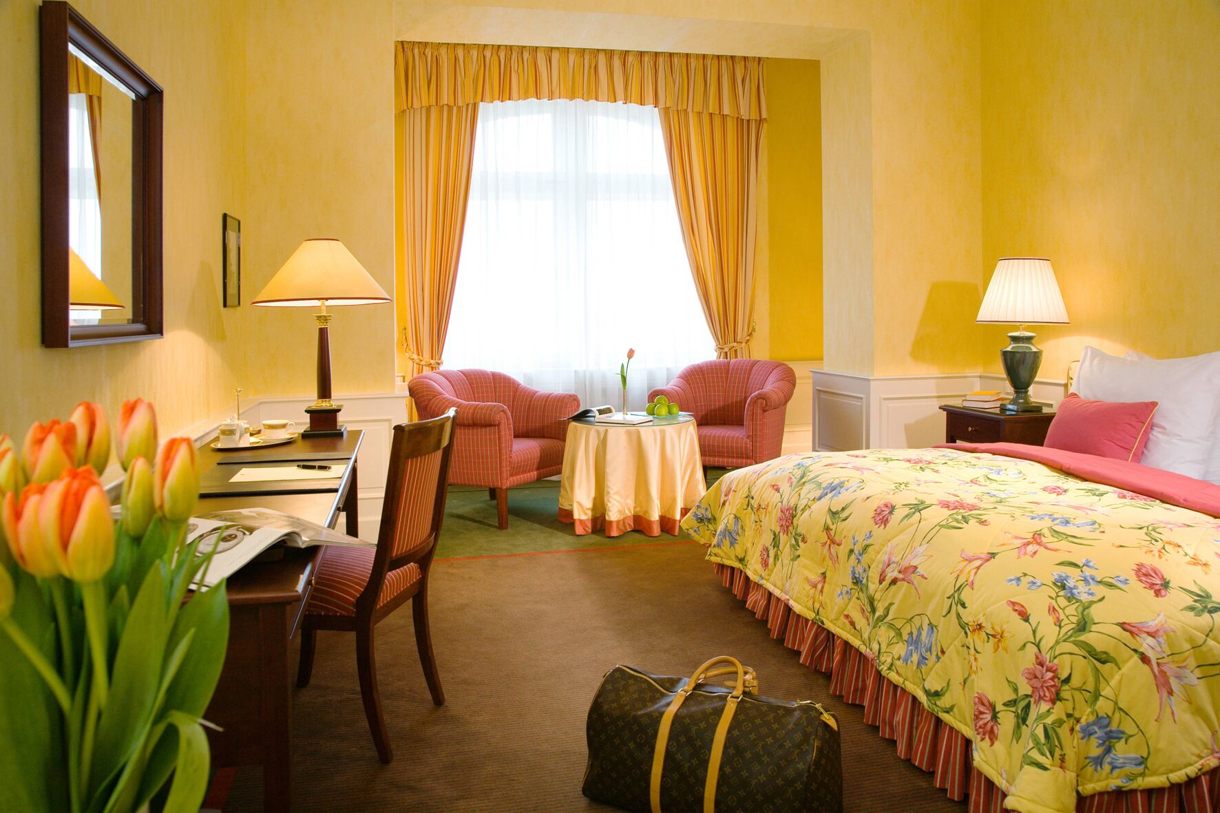 A hotel room with a bed, two chairs, a desk, a lamp, a table with a vase of flowers, and a bag on the floor.