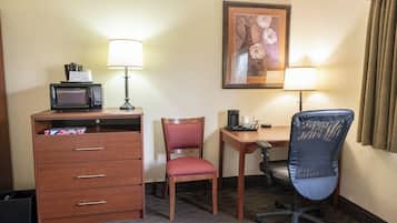 A hotel room with a desk, two chairs, a microwave, a lamp, a framed picture, and a trash bin.