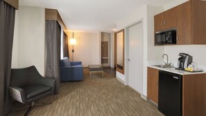 A compact hotel room with a kitchenette, a sofa, a desk, and a lamp.