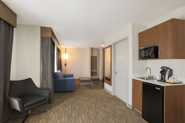 A compact hotel room with a kitchenette, a sofa, a desk, and a lamp.