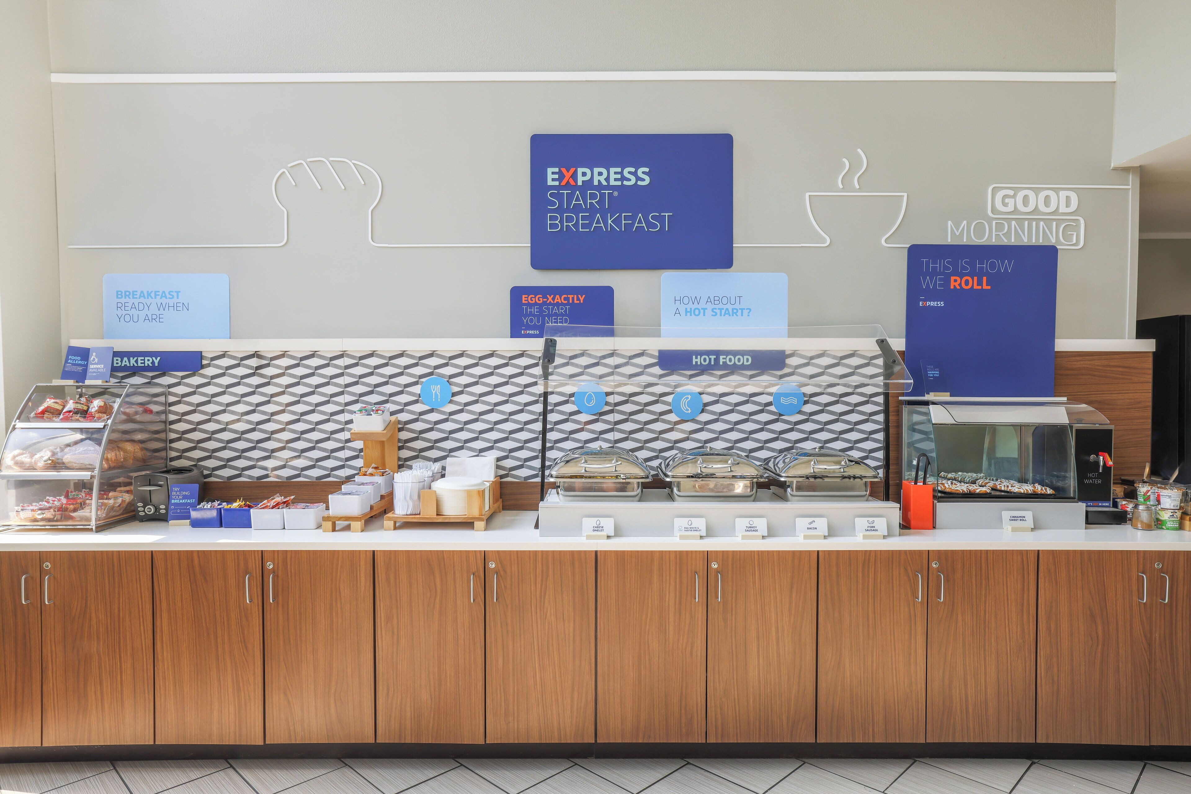 Zona de desayuno tipo buffet con carteles que indican 'EXPRESS START BREAKFAST', 'EGG-XACTLY THE START YOU NEED' y 'COMIDA CALIENTE'.