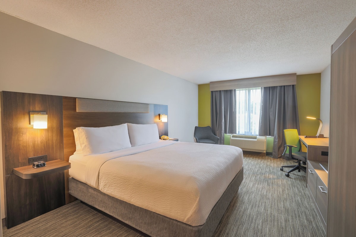 A hotel room with a large bed, a desk with a chair, a nightstand, and a window with curtains.
