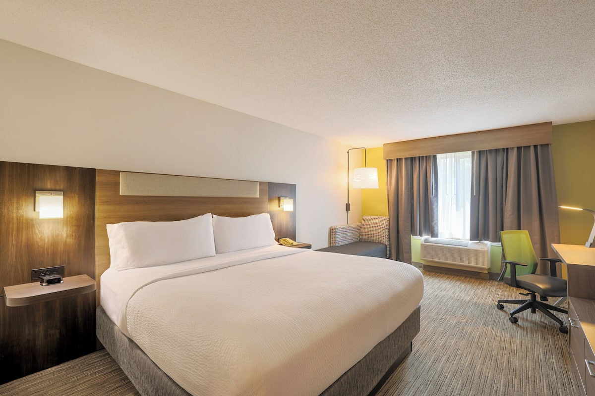 A hotel room with a large bed, a desk, a chair, a lamp, and a window with curtains.