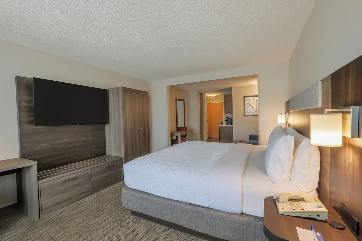 A hotel room with a large bed, a desk with a telephone, a flat-screen TV mounted on the wall, and a mirror above the bed.