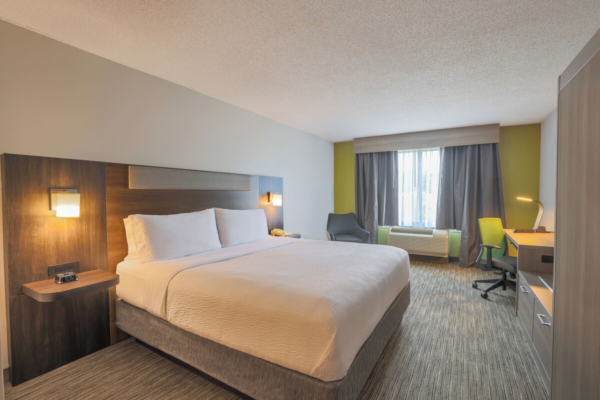 A hotel room with a large bed, a desk with a chair, a nightstand, and a window with curtains.