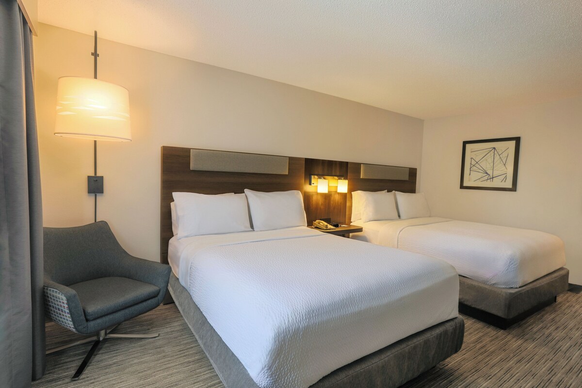 A hotel room with two beds, a chair, a lamp, and a framed picture on the wall.