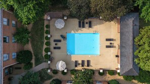 An aerial view of a swimming pool area with lounge chairs and umbrellas, surrounded by greenery and a brick building.