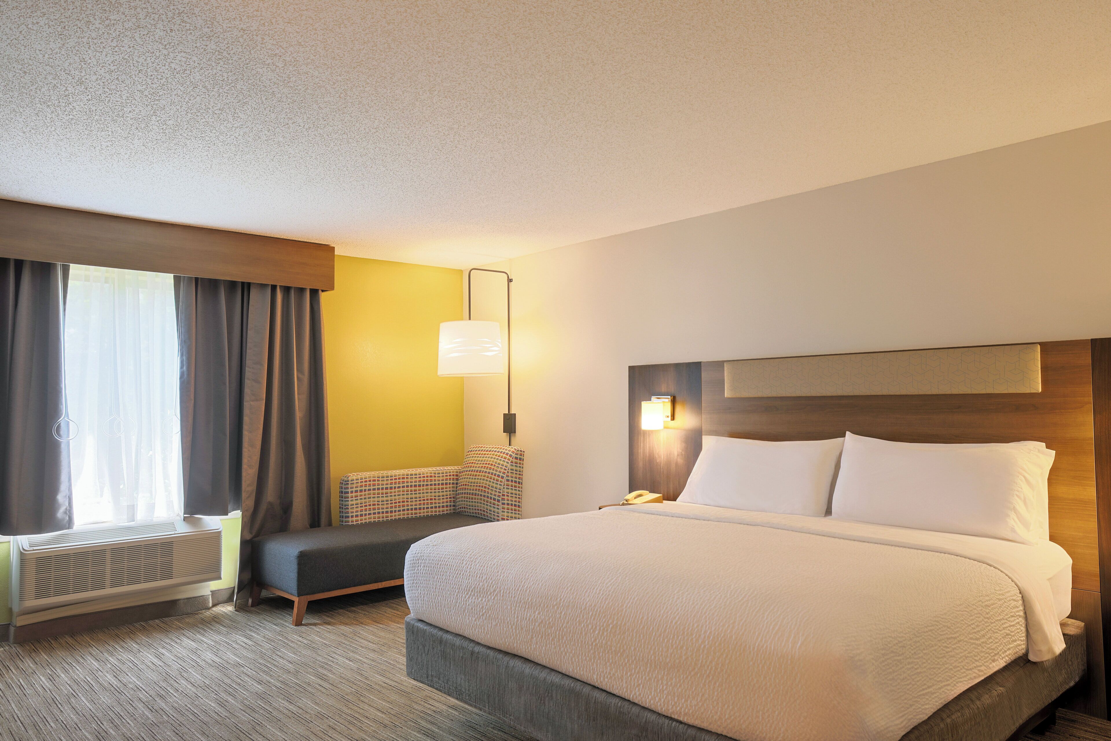 A hotel room with a large bed, a bedside table, a lamp, a chair, and a window with curtains.