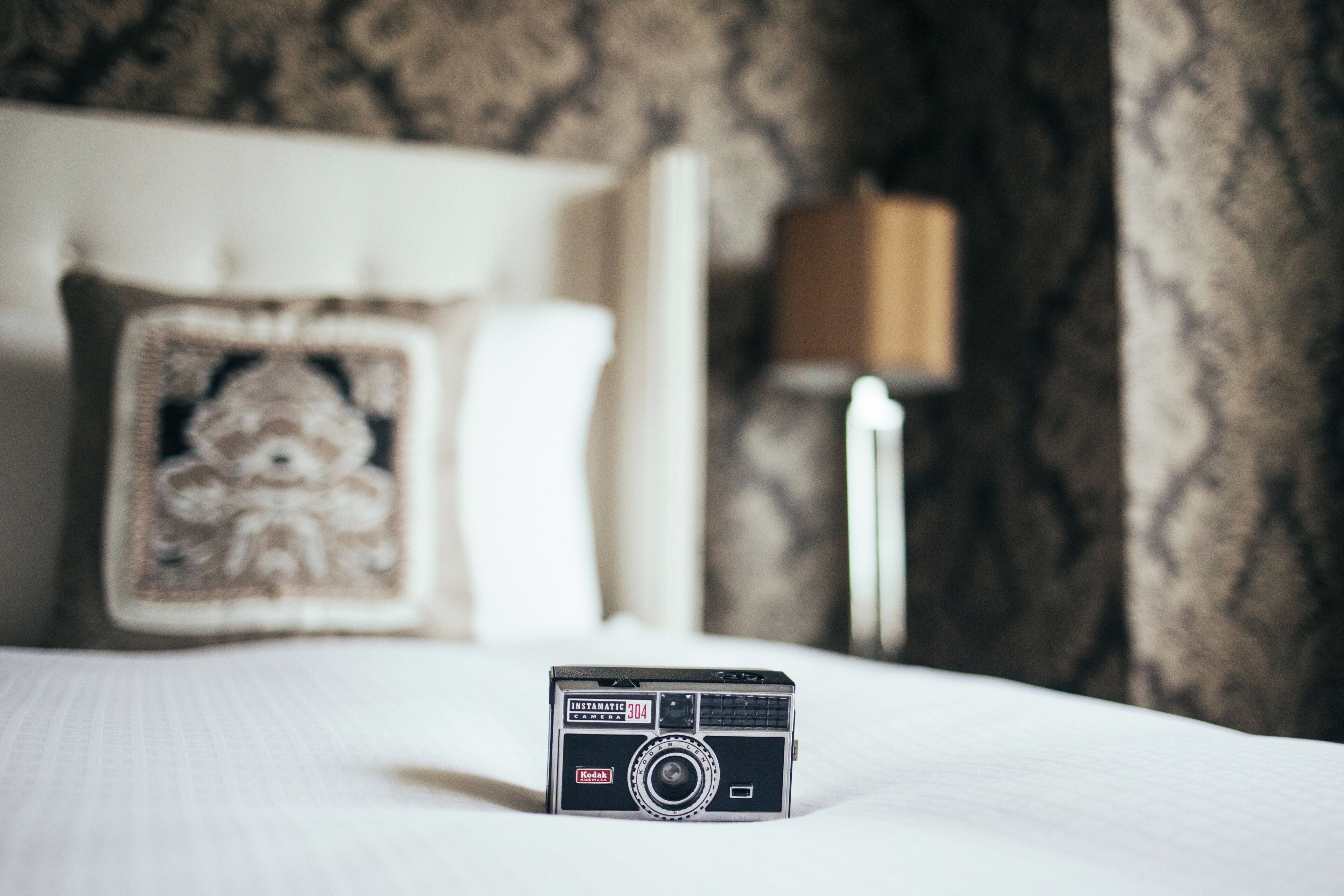 A vintage Kodak Instamatic 404 camera on a bed with a patterned pillow and a lamp in the background.