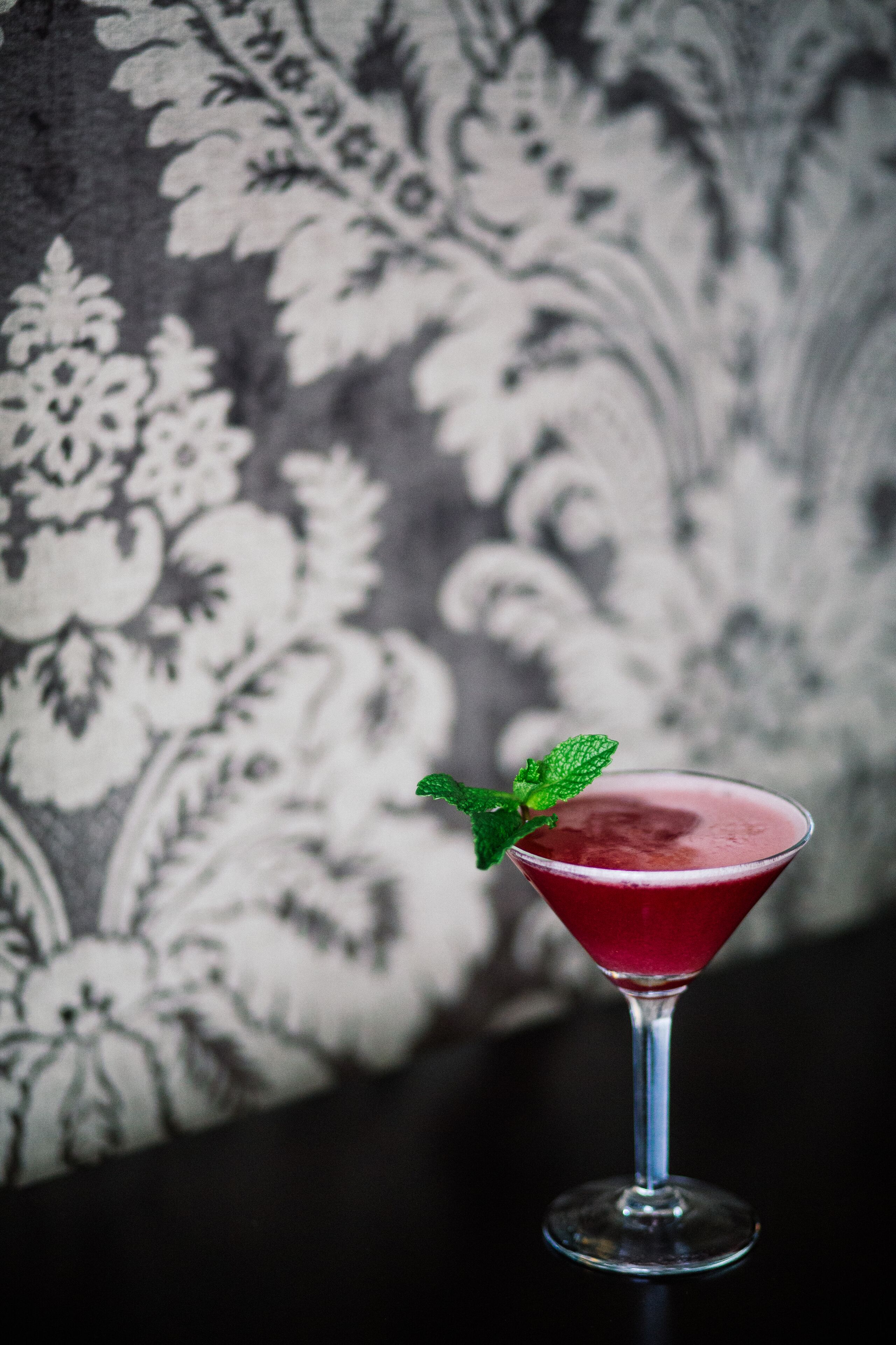 A martini glass with a red drink and a mint garnish on a table.