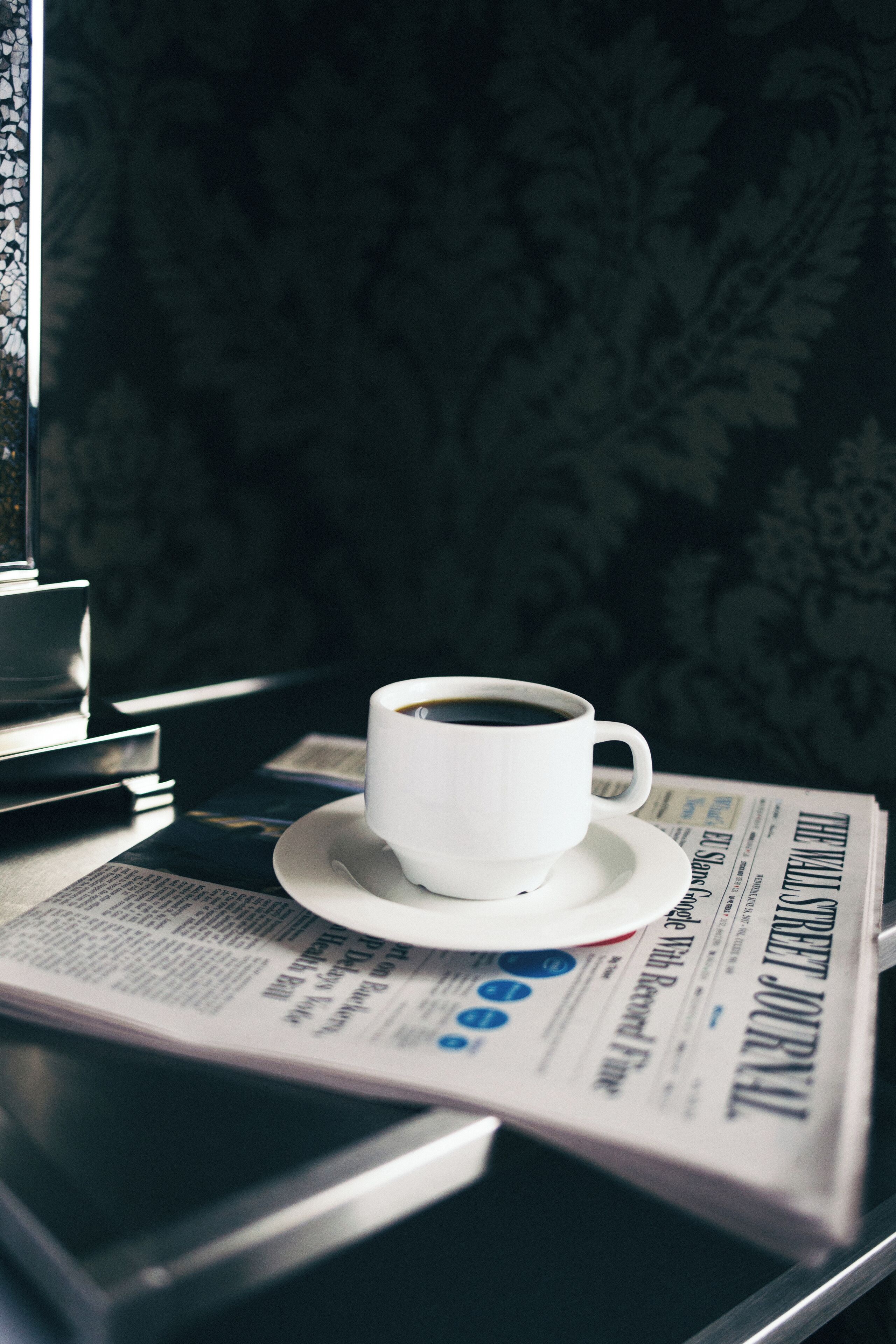 A cup of coffee on a saucer next to a newspaper on a table.