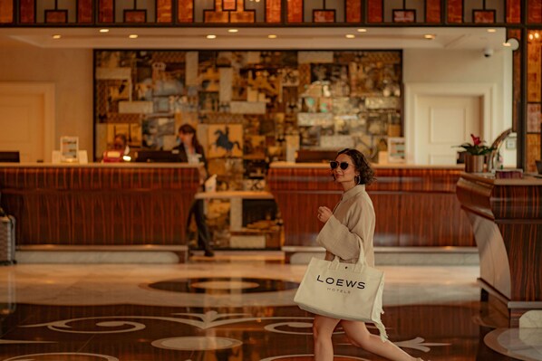 Eine Hotellobby mit einer Rezeption, einer Person, die eine Tasche mit dem Schriftzug 'LOEWS HOTELS' trägt, und einer großen abstrakten Wandkunst.