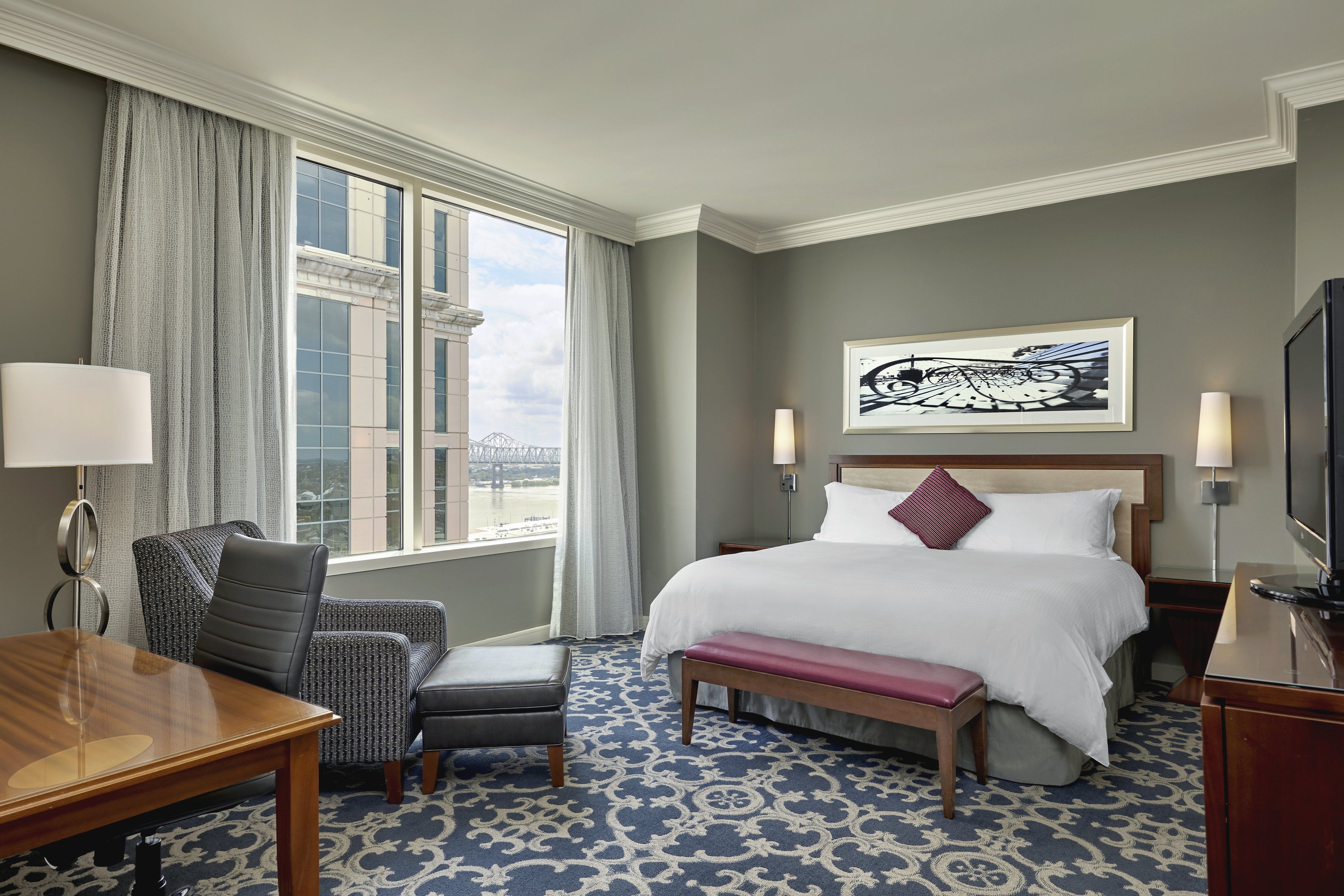 A hotel room with a bed, a desk, a chair, a bench, a TV, and a window with a view of a bridge.