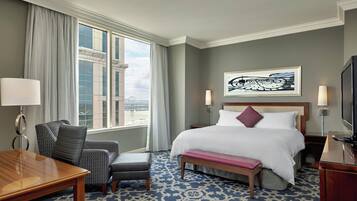 A hotel room with a bed, a desk, a chair, a bench, a TV, and a window with a view of a bridge.