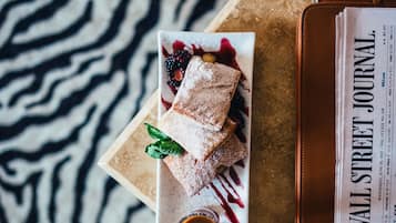 Eine Desserttasse mit Gebäck, Beeren und einem Minzblatt auf einem weißen Teller, begleitet von einer kleinen Soßen-Tasse, platziert auf einem Tisch zusammen mit einer Geldbörse aus Leder und einer Wall Street Journal Zeitung.