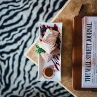 Une assiette à dessert avec une pâtisserie, des baies et une feuille de menthe, le tout disposé sur une assiette blanche, accompagné d’une petite coupelle de sauce, et placé sur une table où se trouvent un portefeuille en cuir et un exemplaire du Wall Street Journal.