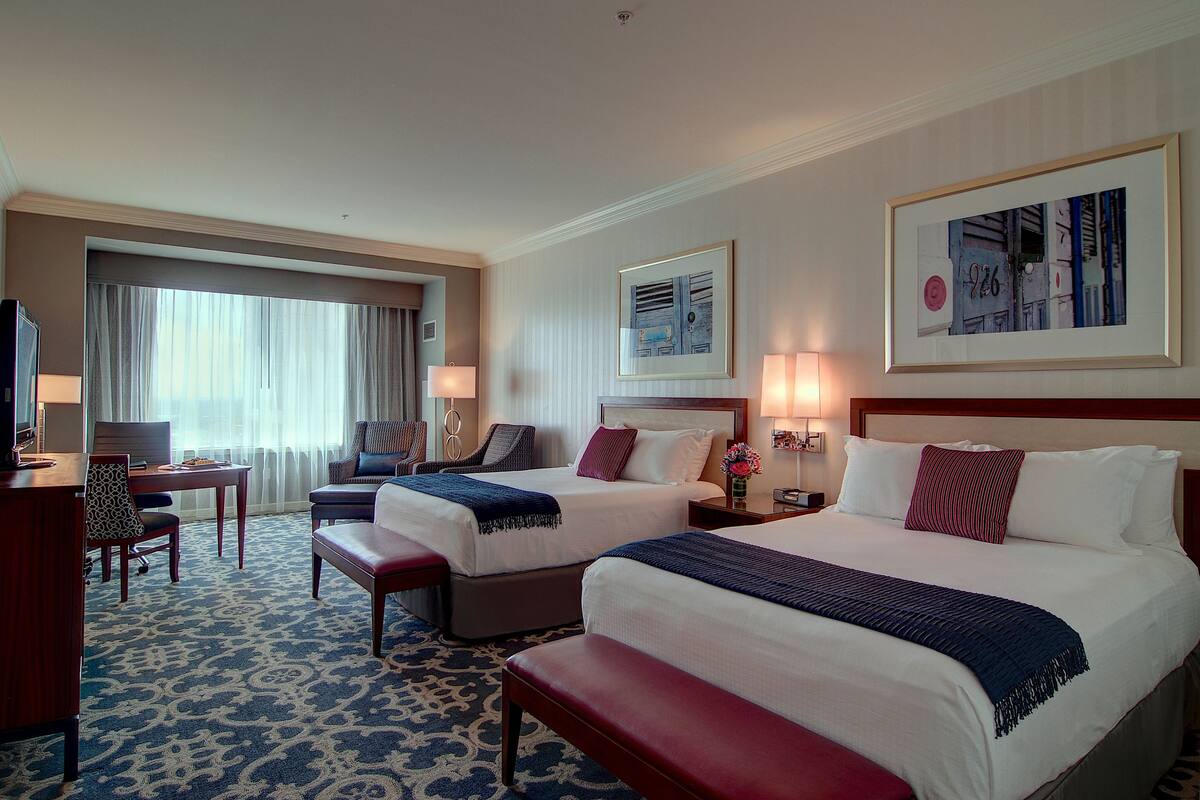 Ein Hotelzimmer mit zwei Betten, einem Schreibtisch, einem Stuhl und einem großen Fenster mit Vorhängen.
