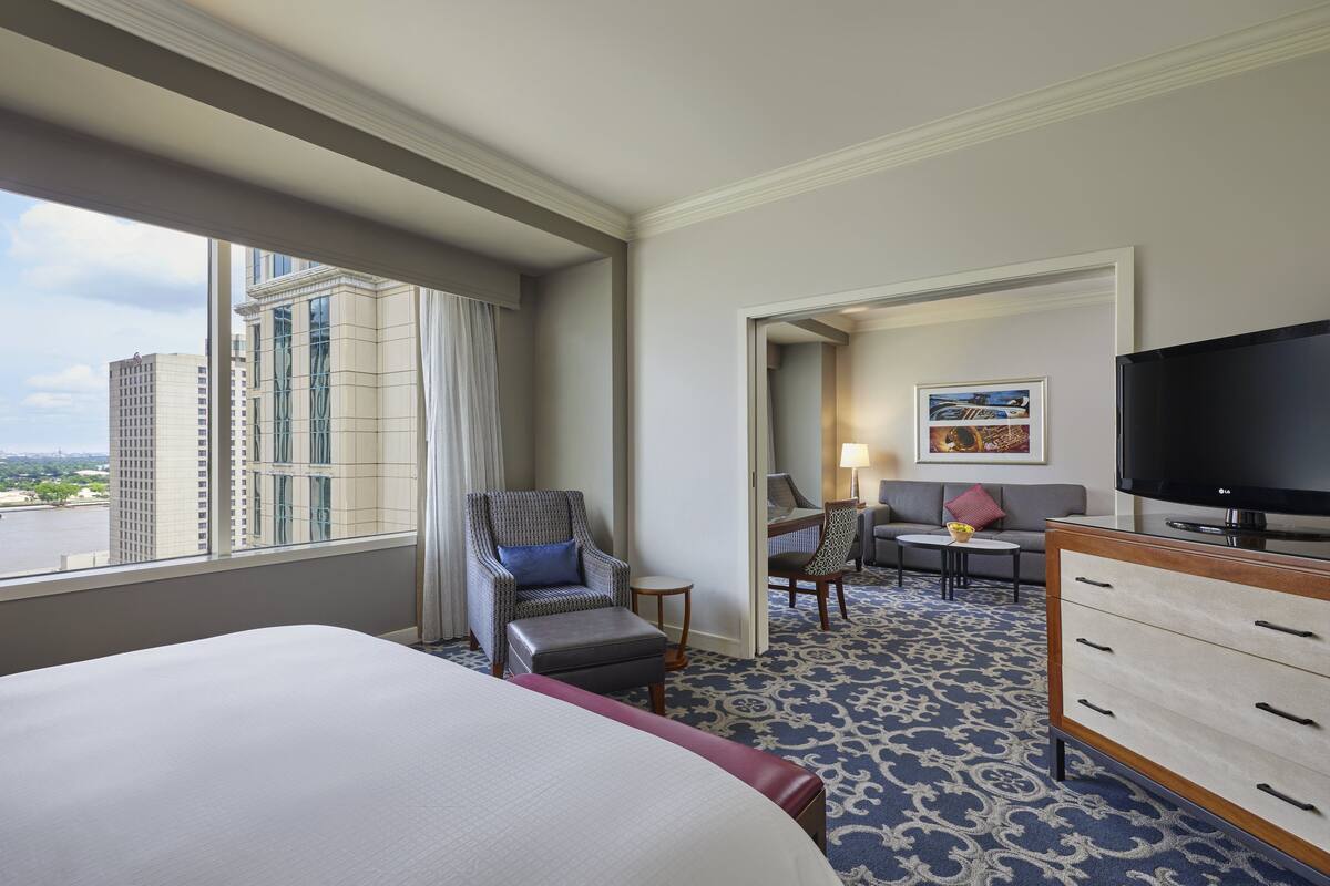 Ein Hotelzimmer mit einem großen Bett, einem Fernseher, einem Schreibtisch mit Stuhl, einer Couch und Blick auf die Stadt durch das Fenster.